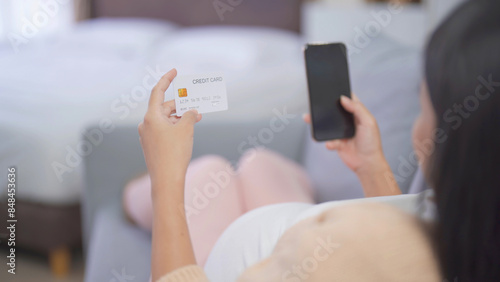 A portrait of happy Asian pregnant woman using a mobile phone device with credit card, shopping online in bedroom at home. Having a baby. Family people lifestyle. Mom love.