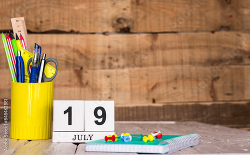 July calendar background with number  19. Stationery pens and pencils in a case on a wooden vintage background. Copy space notepad with pencils and calendar. Planner place for text. photo