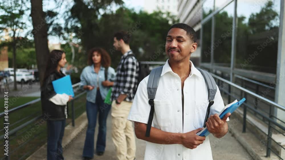 Young African American student smiling with books in the foreground, friends chatting in the background on a university campus. Students socializing outdoors with a focus on a smiling student Young African American student smiling with books in the foreground, friends chatting in the background on a university campus. Students socializing outdoors with a focus on a smiling student
