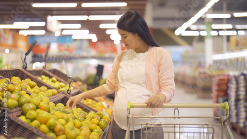 A portrait of happy Asian pregnant woman customer walking along supermarket, buying items on grocery products shelves with basket. Food shopping. Having a baby. Family people lifestyle. Mom.