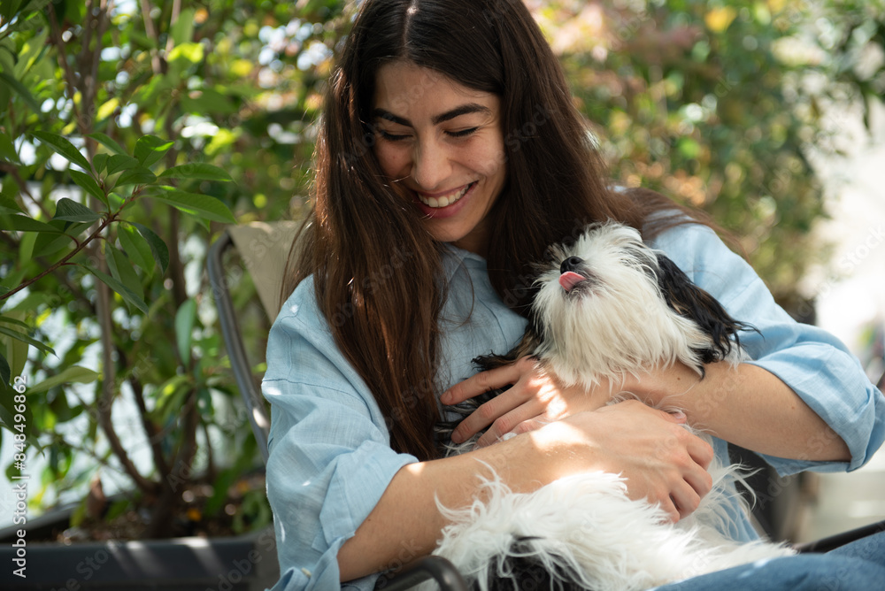 Young woman is smiling while cuddling her adorable black and white dog on a terrace surrounded by plants Young woman is smiling while cuddling her adorable black and white dog on a terrace surrounded by plants