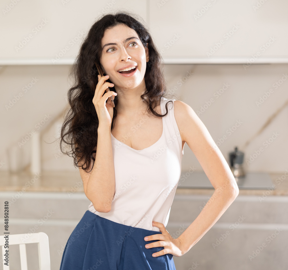 Fototapeta premium Young positive woman chatting on phone in kitchen at home