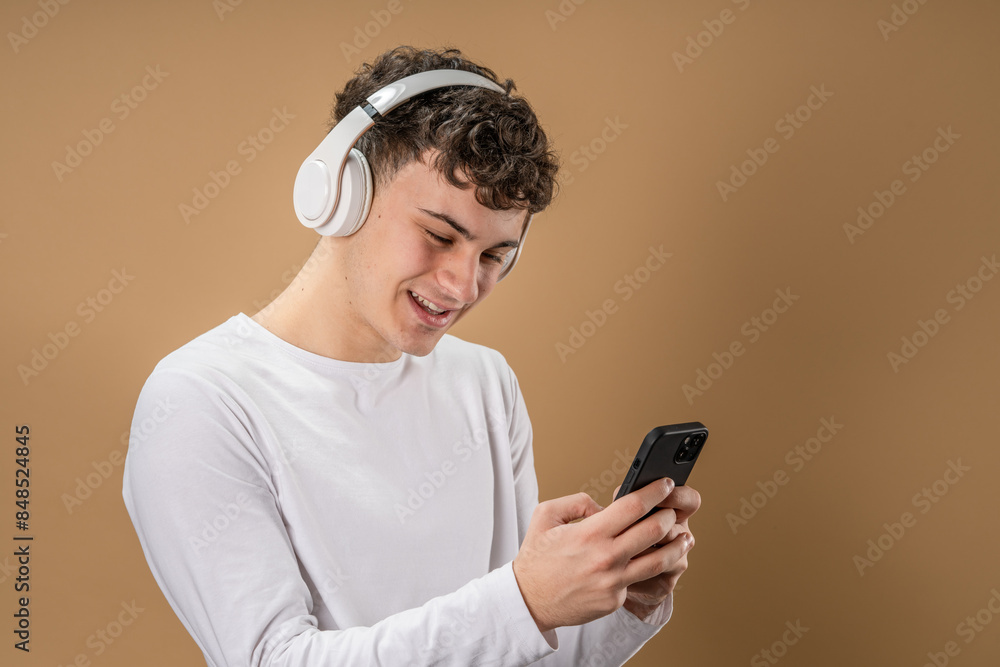 One man caucasian young male stand studio shot on beige background use mobile phone smartphone with headphones listen music send messages texting or browse internet online app copy space One man caucasian young male stand studio shot on beige background use mobile phone smartphone with headphones listen music send messages texting or browse internet online app copy space