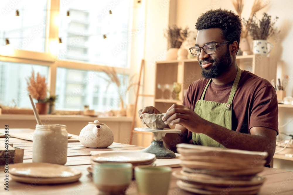 Curly-haired man sitting at the table, working with clay and looking involved Curly-haired man sitting at the table, working with clay and looking involved