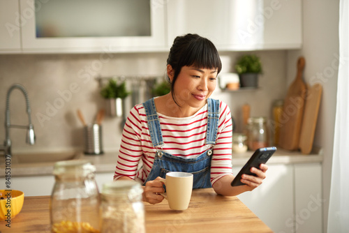 Beautiful Asian woman using mobile phone while drinking coffee in the kitchen