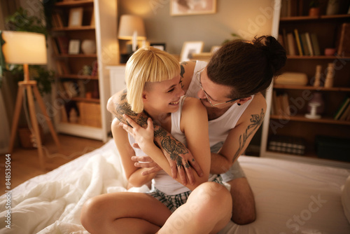 Tablou pe pânză Tattooed man and beautiful woman embracing at home in bed