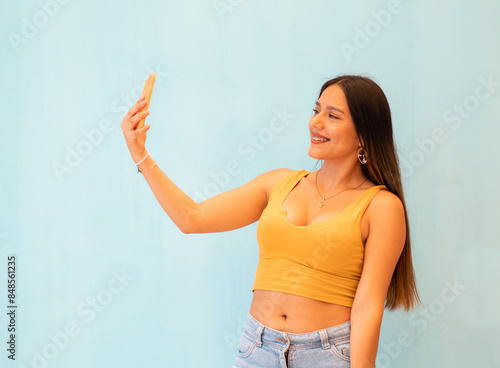 Hermosa joven mujer con polera top amarillo feliz mientras usa un teléfono inteligente y aislada en un fondo gris,Mujer joven tomando selfie en su teléfono 