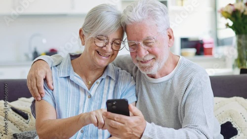 Happy mature family, wife and husband using phone together at home, smiling middle aged man and woman sitting on couch, using mobile device apps, watching video in social network, surfing internet
