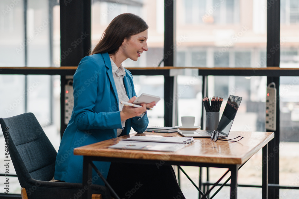 Businesswoman in blue blazer working on a laptop at a wooden desk in a modern office with large windows and natural light. Businesswoman in blue blazer working on a laptop at a wooden desk in a modern office with large windows and natural light.
