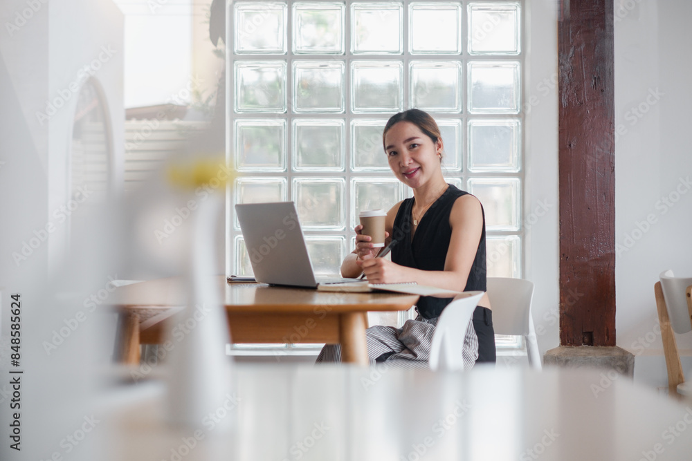 Young woman working on a laptop in a modern cafe, holding a coffee cup and smiling. Bright interior with a glass block wall. Young woman working on a laptop in a modern cafe, holding a coffee cup and smiling. Bright interior with a glass block wall.