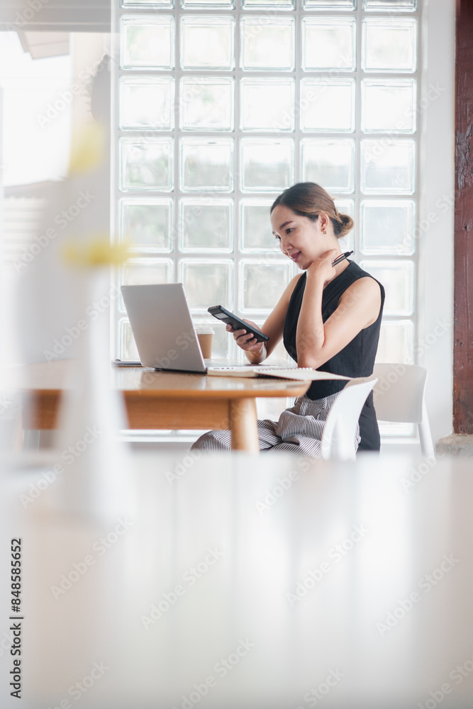 A woman is sitting at a table with a laptop and a cell phone. She is looking at the cell phone and she is focused on something A woman is sitting at a table with a laptop and a cell phone. She is looking at the cell phone and she is focused on something