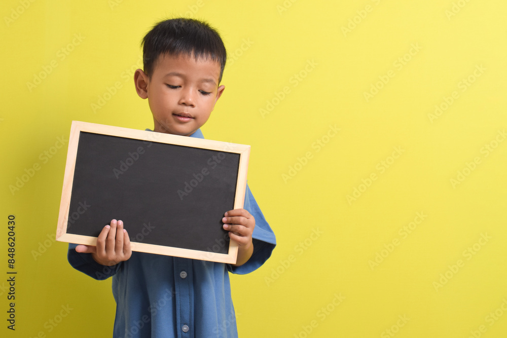 Cute little Asian boy holding a blank chalk board isolated on yellow background. Space for text, copy space, mockup banner Cute little Asian boy holding a blank chalk board isolated on yellow background. Space for text, copy space, mockup banner