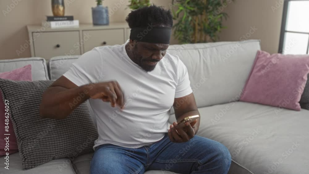 A young, handsome african american man with a beard interacts with his smartphone while sitting on a comfortable couch in his cozy living room. A young, handsome african american man with a beard interacts with his smartphone while sitting on a comfortable couch in his cozy living room.