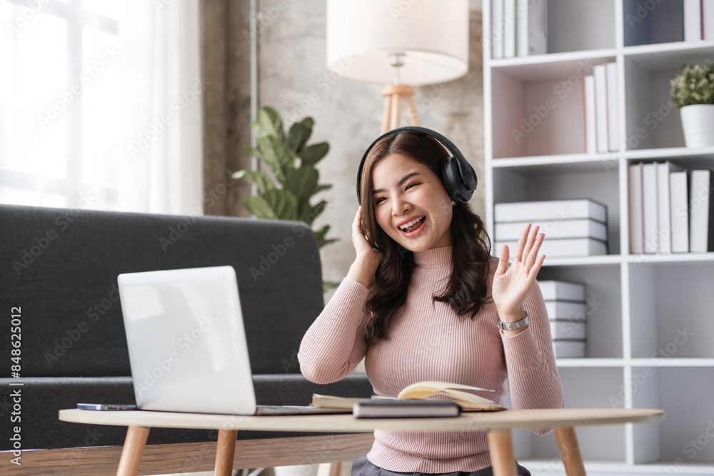 Young Woman Sitting and Working in Modern Living Room with Laptop, Headphones, and Books, Smiling and Waving, Cozy Home Office Setup, Productive and Comfortable Work Environment Young Woman Sitting and Working in Modern Living Room with Laptop, Headphones, and Books, Smiling and Waving, Cozy Home Office Setup, Productive and Comfortable Work Environment