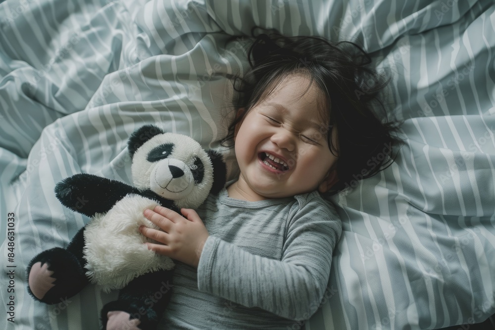 In the top view, an Asian child is lying on the bed, playing with a panda plush toy and smiling happily In the top view, an Asian child is lying on the bed, playing with a panda plush toy and smiling happily