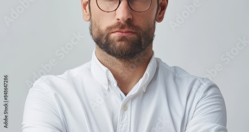 A man with a beard wearing glasses and a white shirt stares intensely at the camera with arms crossed. photo