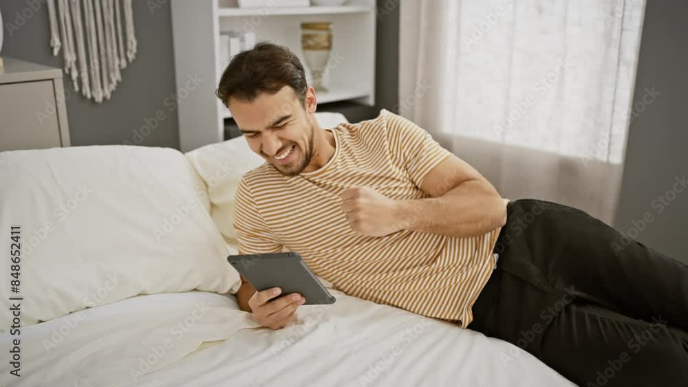 A cheerful hispanic man with a beard comfortably lounges in bed using a tablet in a modern bedroom. A cheerful hispanic man with a beard comfortably lounges in bed using a tablet in a modern bedroom.