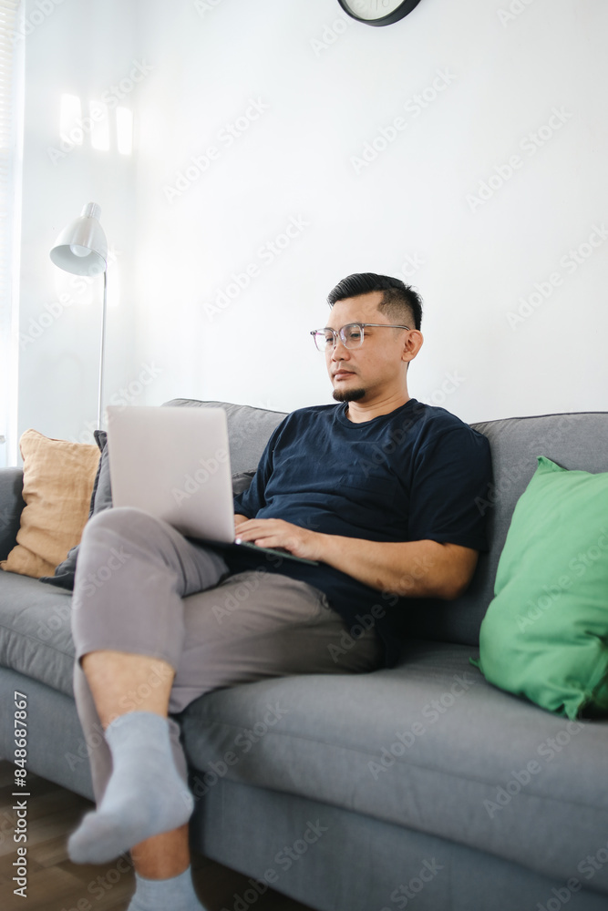 Asian Man Working On Laptop Sitting On Sofa At Home. Remote job concept. Asian Man Working On Laptop Sitting On Sofa At Home. Remote job concept.