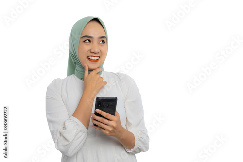 Beautiful young Asian woman in green hijab and white blouse holding smartphone and touching chin, smiling curiously thinking about something isolated on white background © Sewupari Studio