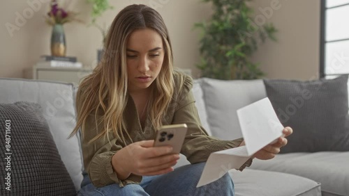 A young woman sits indoors on a couch, looking concerned while holding a smartphone and a piece of paper, creating a relatable image of everyday stress and financial worries.