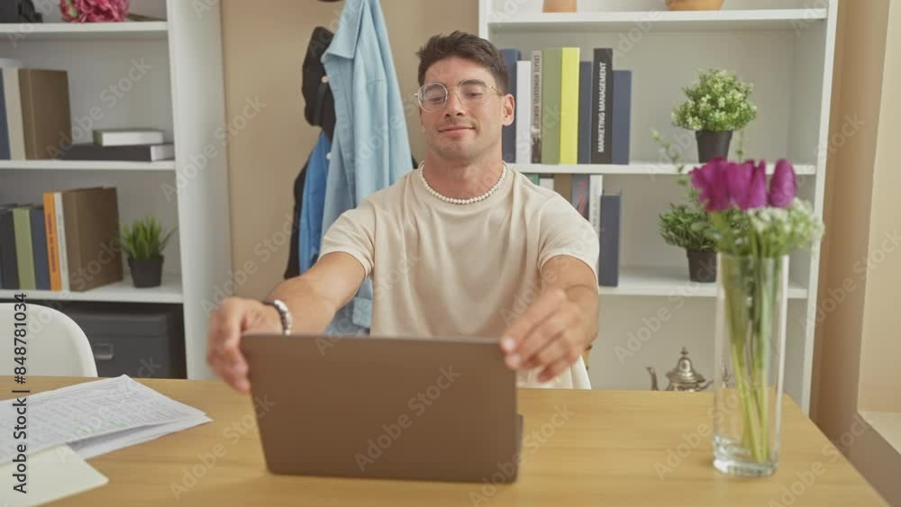 A cheerful young hispanic man closes a laptop in a modern home office with shelves and flowers. A cheerful young hispanic man closes a laptop in a modern home office with shelves and flowers.