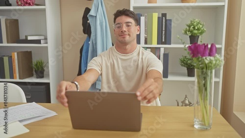 A cheerful young hispanic man closes a laptop in a modern home office with shelves and flowers. photo