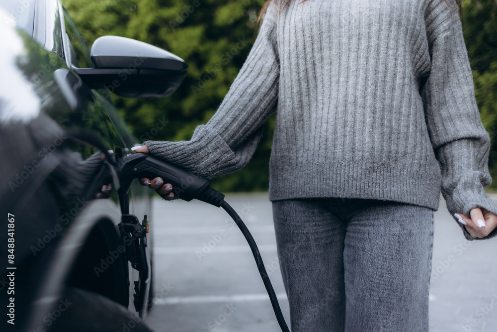Obraz premium Woman plugging in the charger into a socket of her blue electric car at a charging station in the street