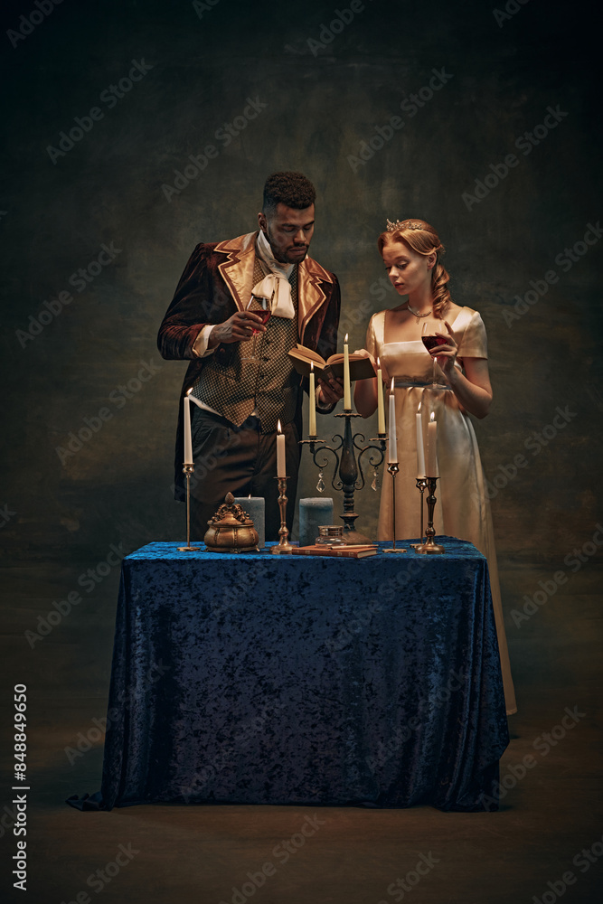 Cozy evening atmosphere. Elegant young woman and handsome man, royal family standing by the table with candles, drinking wine and reading books. History, retro and vintage, comparison of eras concept Cozy evening atmosphere. Elegant young woman and handsome man, royal family standing by the table with candles, drinking wine and reading books. History, retro and vintage, comparison of eras concept