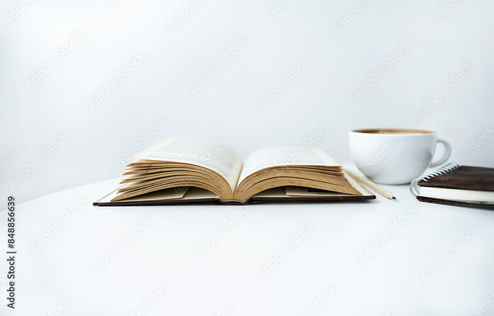 Close-up shot, white coffee cup and note book with open book on white table and white wall on background Close-up shot, white coffee cup and note book with open book on white table and white wall on background