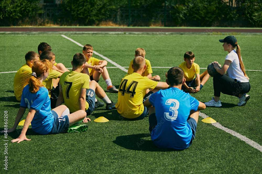 Obraz premium Active Youth Engagement. Children sitting on grass and listening to female football coach instructions, learning soccer game strategies and skills. Sport, school, childhood, active lifestyle concept