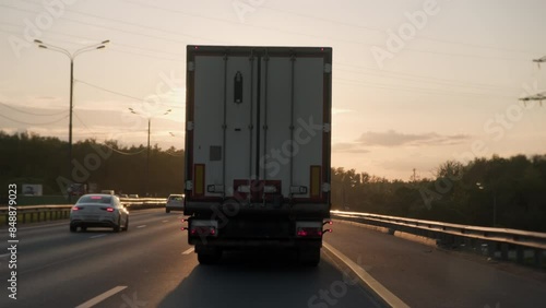 White cargo truck van with freight semi-trailer driving along highway in evening time at sunset, rear view.