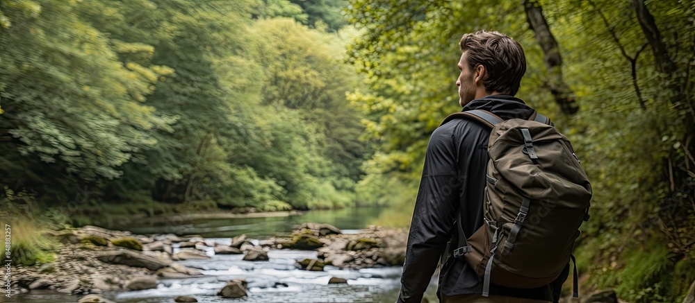 A young man with a backpack is seen from behind exploring a forest by a river The image shows him engaging in active hiking with tourist gear embodying an active lifestyle. Copy space image A young man with a backpack is seen from behind exploring a forest by a river The image shows him engaging in active hiking with tourist gear embodying an active lifestyle. Copy space image