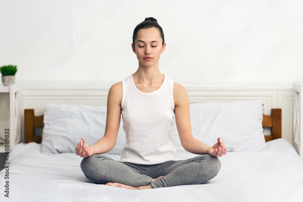 A young woman sits in a meditative pose on a bed, eyes closed, in a bright, well-lit room. A young woman sits in a meditative pose on a bed, eyes closed, in a bright, well-lit room.