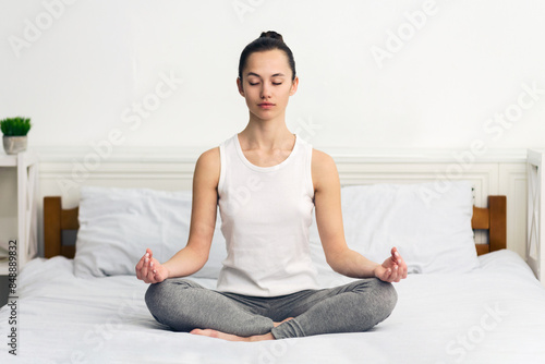 A young woman sits in a meditative pose on a bed, eyes closed, in a bright, well-lit room.