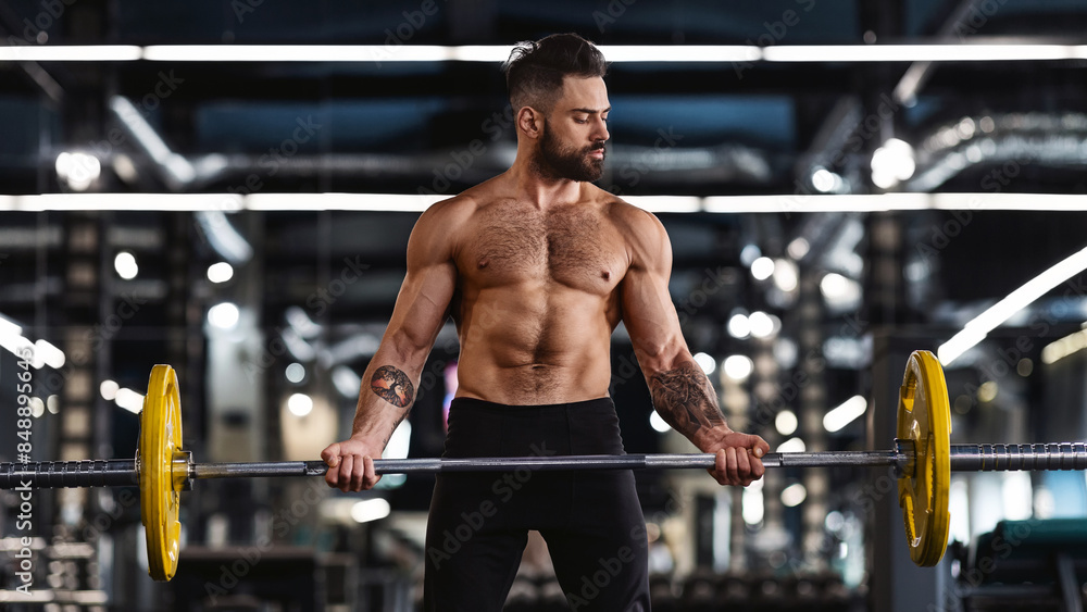 A muscular man lifts weights in a gym. He is shirtless and wearing black pants. The gym is brightly lit and has a lot of equipment.