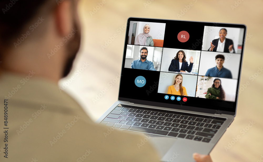 Man using a laptop while a group of people are visible on the screen. The person is engaged in virtual communication or collaboration with the individuals displayed on the laptop screen. Man using a laptop while a group of people are visible on the screen. The person is engaged in virtual communication or collaboration with the individuals displayed on the laptop screen.