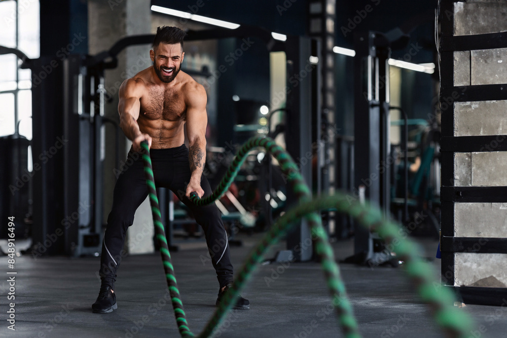 Fototapeta premium Crossfit training. Naked muscular man exercising with battle ropes at gym, free space