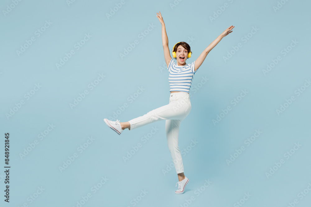 Full body side view young excited fun happy woman wears striped t-shirt casual clothes listen to music in headphones isolated on plain pastel light blue background studio portrait. Lifestyle concept. Full body side view young excited fun happy woman wears striped t-shirt casual clothes listen to music in headphones isolated on plain pastel light blue background studio portrait. Lifestyle concept.