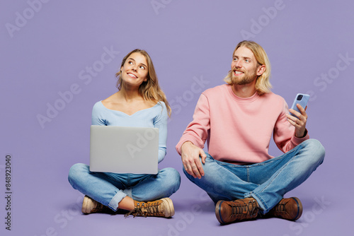 Full body young couple two friends family IT man woman wear pink blue casual clothes together sitting use mobile cell phone work on laptop pc computer look aside isolated on plain purple background.