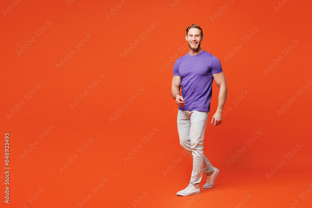 Fototapeta premium Full body young smiling cheerful fun happy Caucasian man he wear purple t-shirt casual clothes walking going looking camera isolated on red orange wall background studio portrait. Lifestyle concept