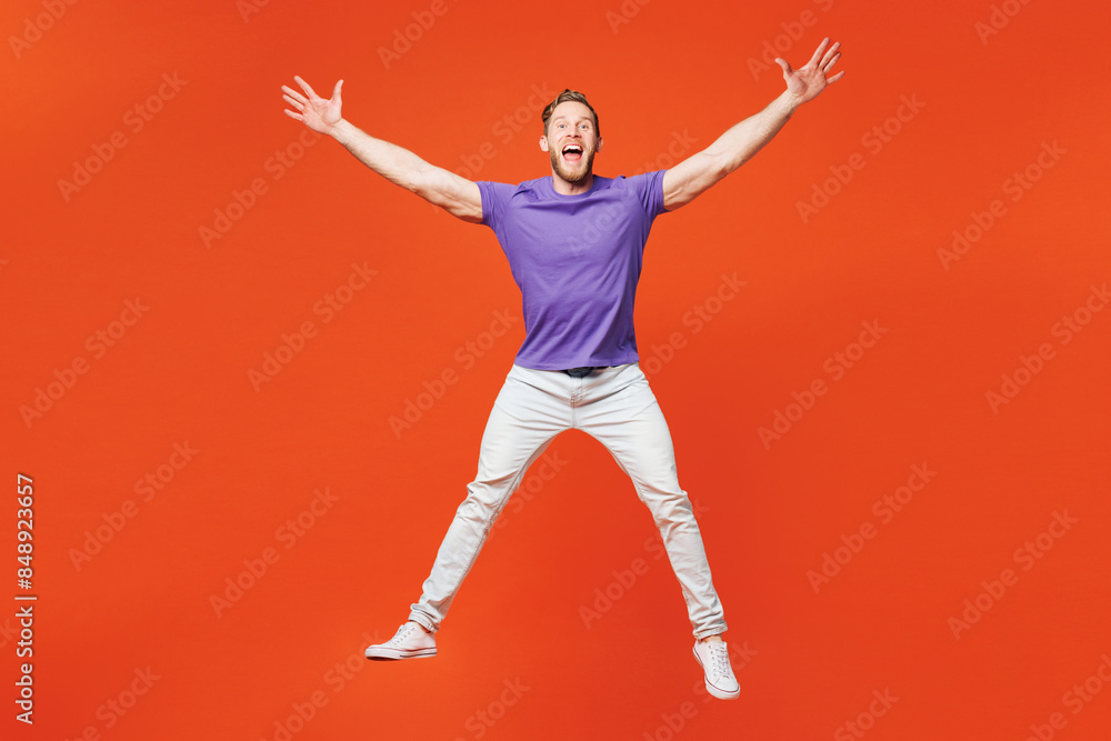 Full body young smiling happy fun man he wearing purple t-shirt casual clothes jump high with outstretched hands legs look camera isolated on red orange background studio portrait. Lifestyle concept.