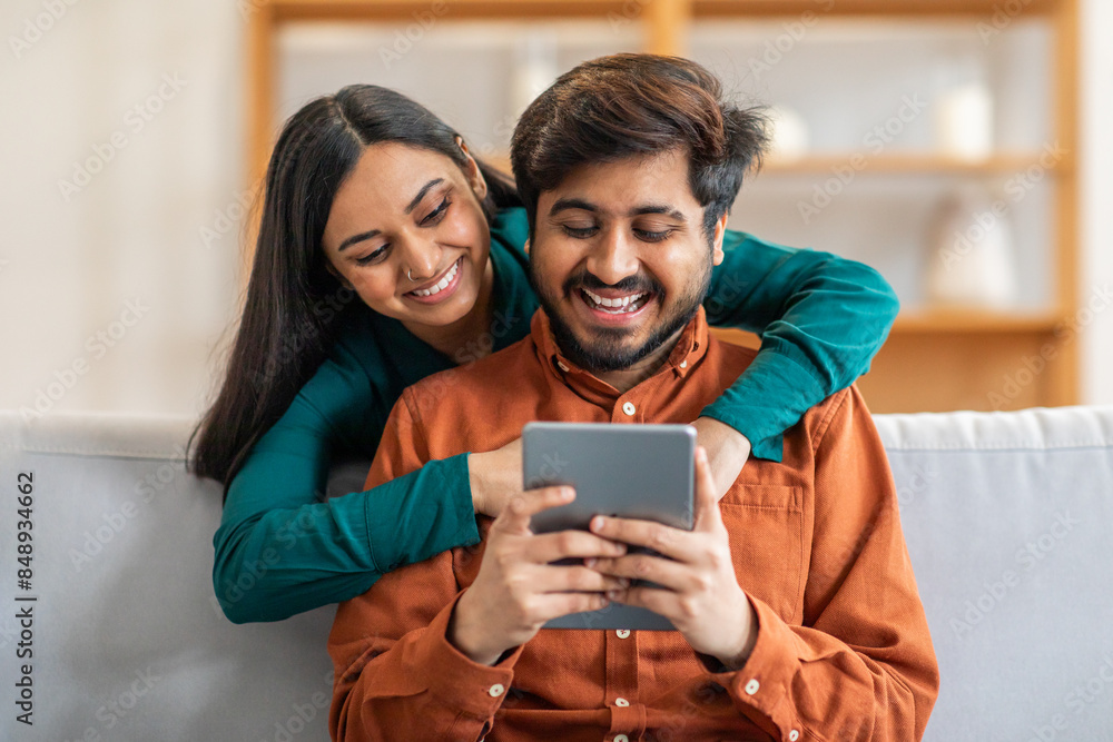 Indian man and woman are seated on a couch, both focused on a tablet screen. They appear engaged as they interact with the device, possibly watching a video or scrolling through information Indian man and woman are seated on a couch, both focused on a tablet screen. They appear engaged as they interact with the device, possibly watching a video or scrolling through information