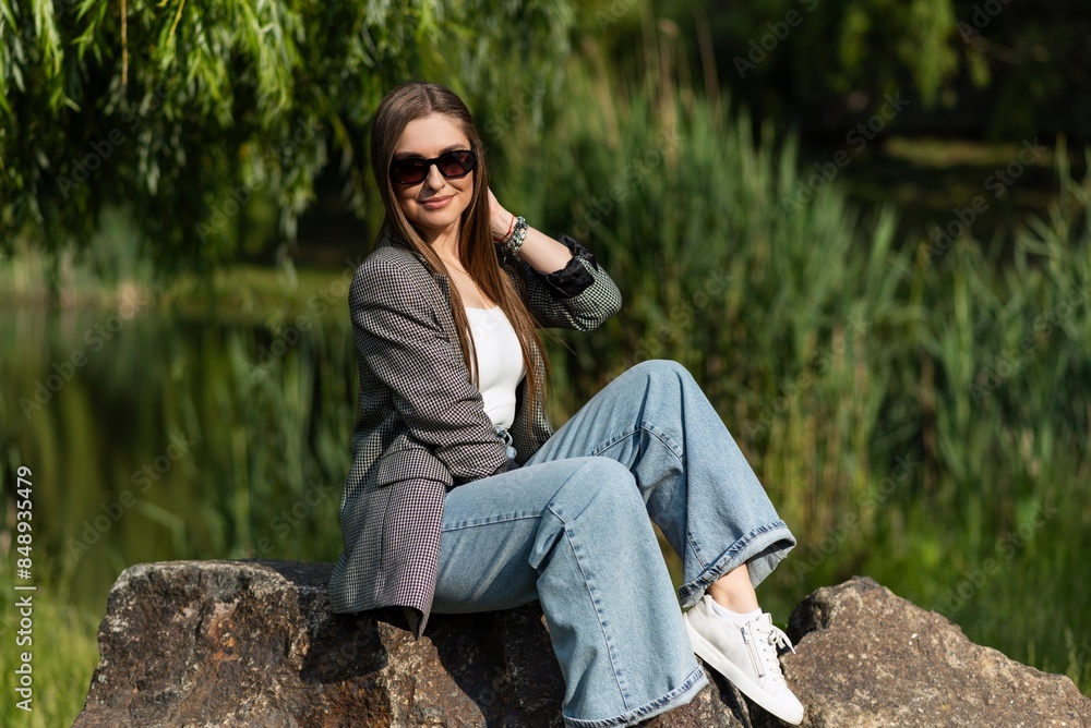 Fototapeta premium Attractive young woman in sunglasses and stylish casual clothes with pleasure posing sitting on a big stone in the park while walking in the park on a spring day. Relax, freedom.