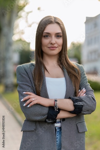 Portrait of attractive Caucasian young woman businesswoman in formal casual clothes with crossed arms, confidently looking at camera while walking outside on sunny warm spring day. photo