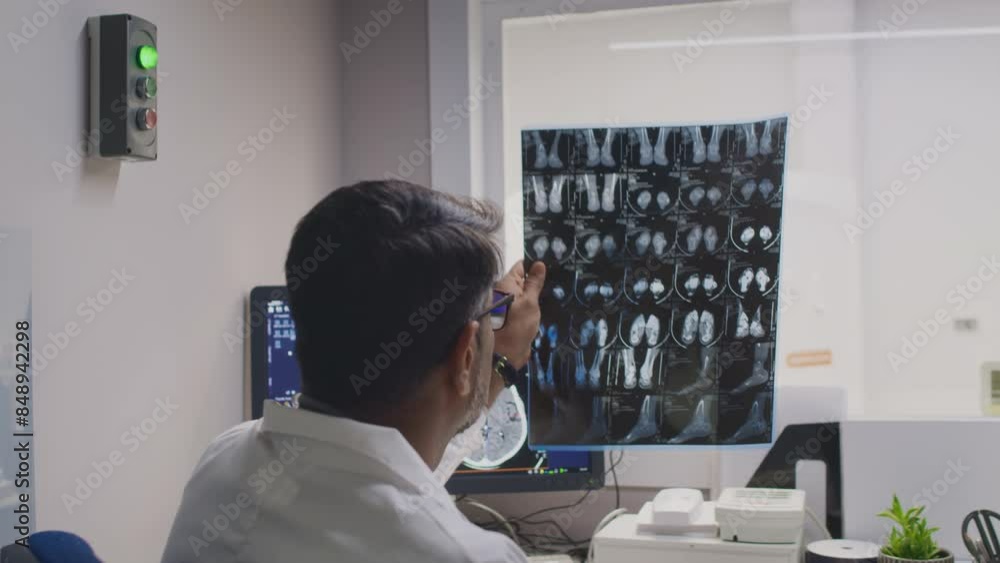 Medium slowmo back view of mature male radiologist in white coat sitting at table and writing medical report, holding up abnormal feet x-ray on film against light in clinic room Medium slowmo back view of mature male radiologist in white coat sitting at table and writing medical report, holding up abnormal feet x-ray on film against light in clinic room