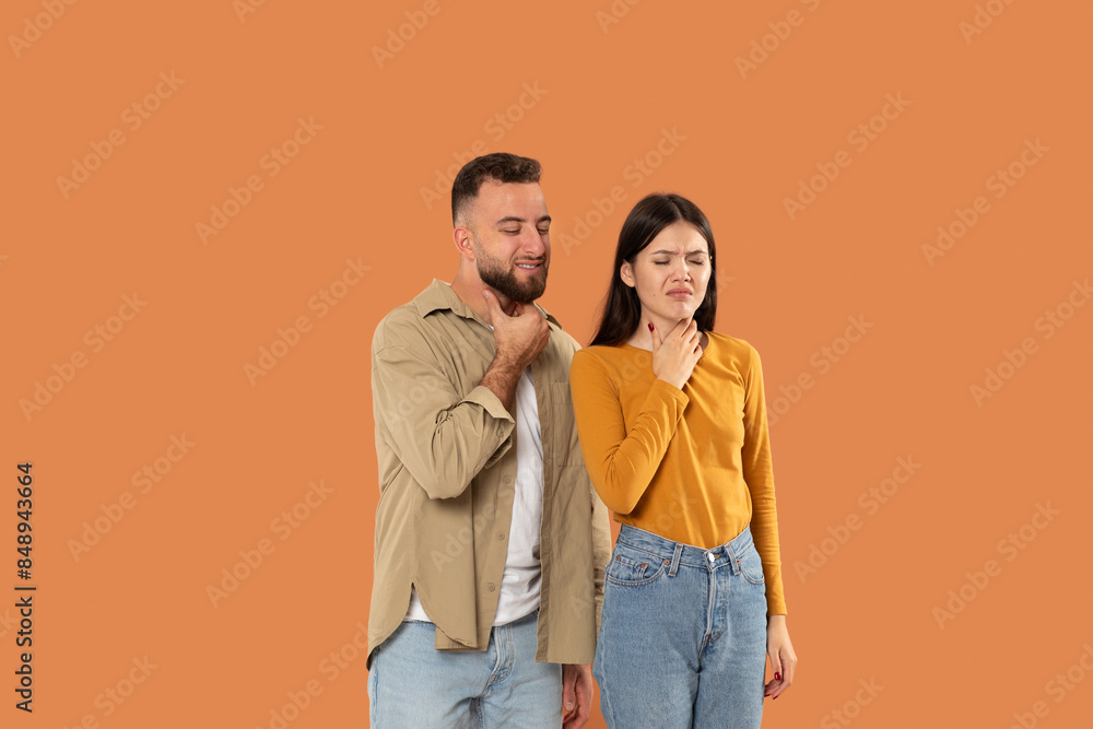 A young couple, casually dressed, stands against an orange background. They both hold their necks and display expressions of discomfort or pain. A young couple, casually dressed, stands against an orange background. They both hold their necks and display expressions of discomfort or pain.