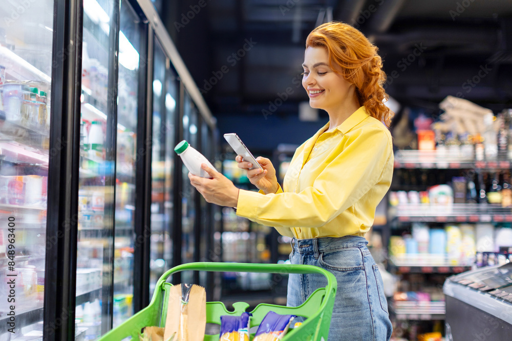 Fototapeta premium Cheerful European woman scanning qr code of daily product using smartphone, standing near refrigerator. Concept of shopping and consumerism.