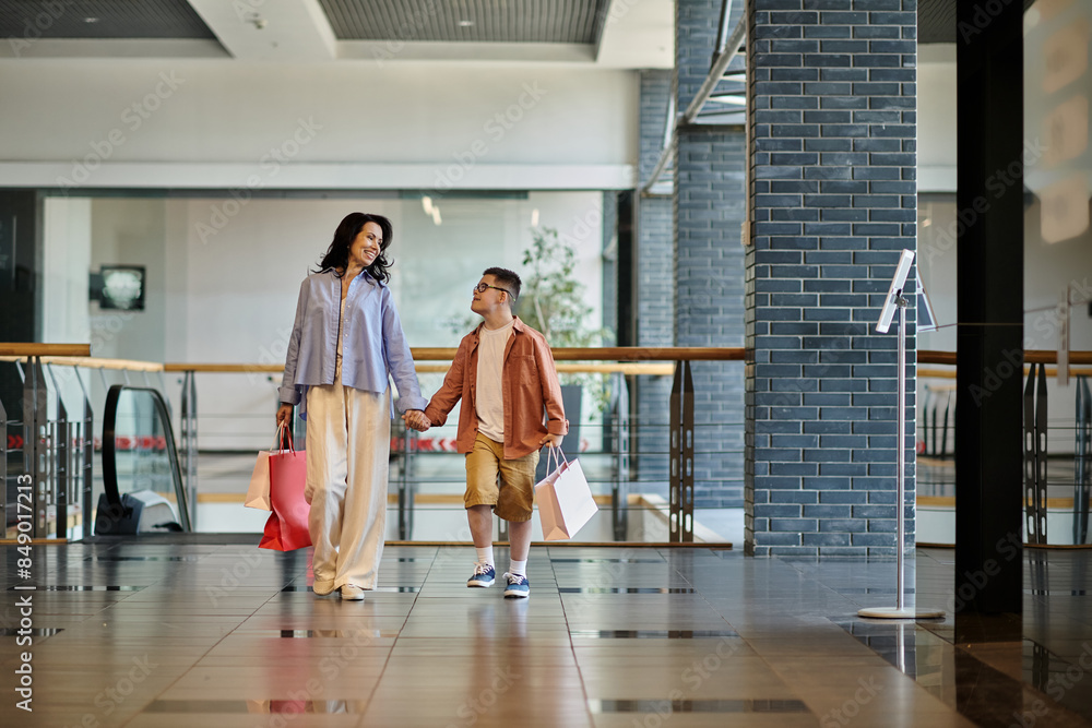 Fototapeta premium A mother and her son with Down syndrome walk hand-in-hand through a shopping mall, carrying shopping bags.