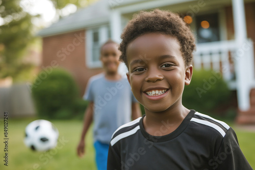 Cheerful African American boy with a soccer ball standing in front of a house with another child in the background. © Irina B