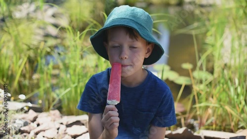 Cute pre-teen boy in green panama eating ice cream against the background of a decorative lake with reeds in the yard photo